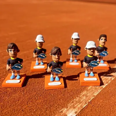 Trofeos de un torneo PELOTEO, con forma de jugadores, apoyados sobre una cancha de polvo de ladrillo.
